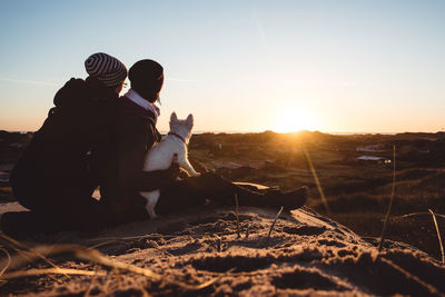 Dog sitting and two people on landscape against clear sky during sunset