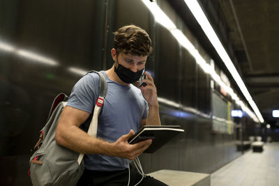 Young man wearing protective face mask talking on phone while holding digital tablet at subway during pandemic