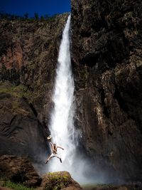 Happy young man with hat jumping against waterfall