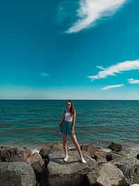 Full length of young woman standing on rocks by sea against sky