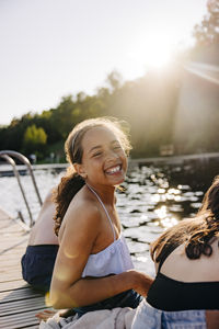 Side view portrait of happy girl laughing while sitting with friends on jetty near lake