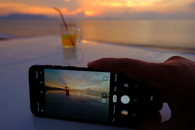 Cropped image of hand holding smart phone against sea during sunset