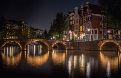 Illuminated bridge over river by buildings in city at night