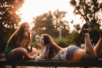Young woman blowing bubbles while her girlfriend lying on bench
