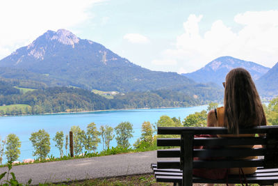 Rear view of woman sitting on bench by lake against mountains