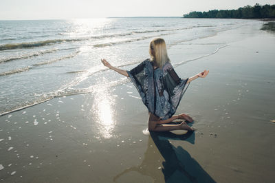 Full length of young woman kneeling with arms outstretched on shore at beach