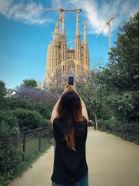 Rear view of woman photographing cathedral through smart phone against sky