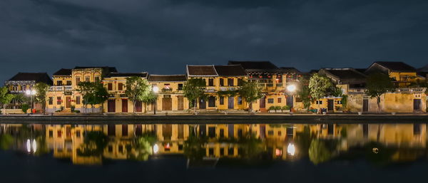 Illuminated buildings by lake against sky at night