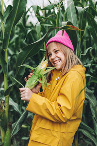 Stylish teenage girl in yellow raincoat and hot pink hat laughing on corn field