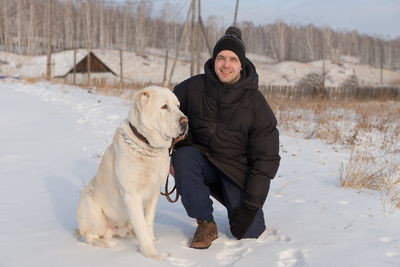 A young man is sitting with a big dog on a rural road against village on a frosty winter day.