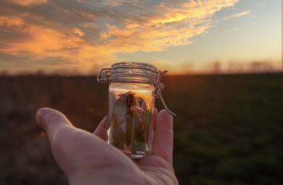 Midsection of person holding glass jar against sky during sunset