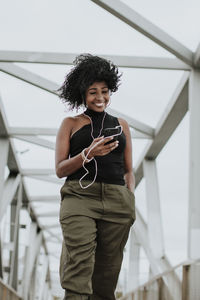 Smiling woman using smart phone listening to music through earphones standing on bridge in city