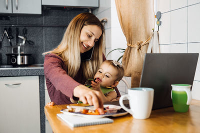 Mother feeding daughter while sitting on table at home