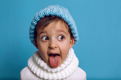 Boy in a blue knitted hat and white sweater shows his tongue on a blue background