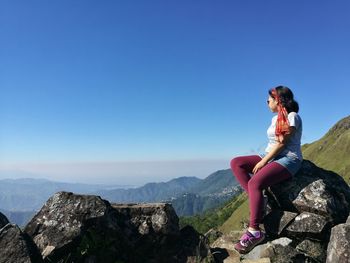 Woman sitting on rock against mountains against clear blue sky