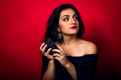 Thoughtful female model holding red grapes while looking away against red background