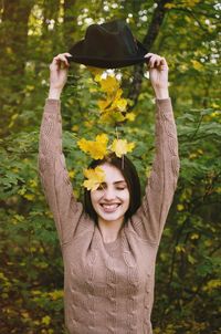 Autumn leaves falling from hat held by cheerful young woman against trees