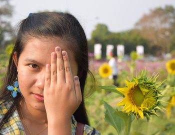 Close-up portrait of young woman covering eye while standing at sunflower field