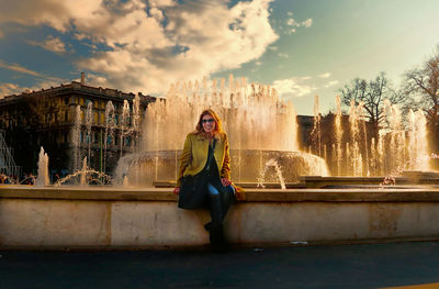 Young woman sitting on retaining wall against fountain during sunset
