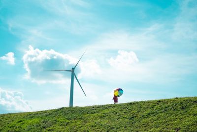 Rear view of man standing on mountain against sky holding rainbow umbrella, beside windmill