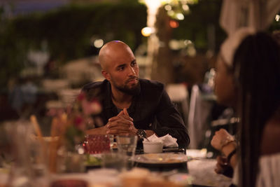 Young man with shaved head looking at female friend while sitting at table during dinner party