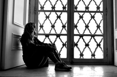 Side view of woman looking through window while sitting on floor