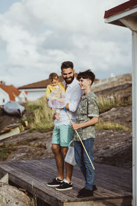 Smiling father looking at son while holding daughter against sky