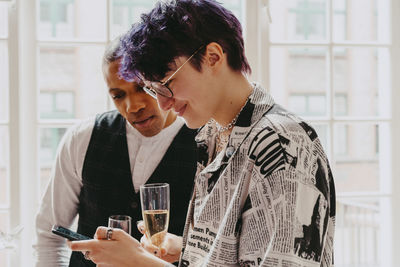 Smiling non-binary person sharing smart phone with male colleague during event at convention center