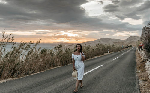 Front view of beautiful young woman in white dress standing on open road.