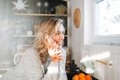 Young woman with blonde curly hair in grey sweater using mobile phone in kitchen at house