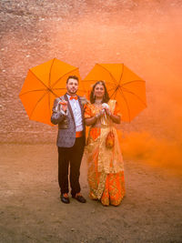 Full length portrait of bride and bridegroom standing with umbrellas on field