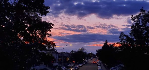 Silhouette trees by street against sky at sunset