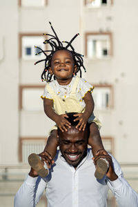 Cheerful african american father in shirt carrying little daughter on shoulders and jumping while spending time together on street in city in sunlight