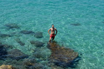 Full length of shirtless young man standing on rock in sea