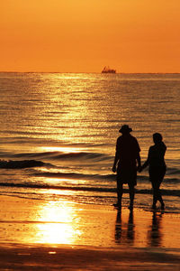 Silhouette couple holding hands while walking at beach against clear sky during sunset