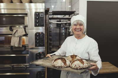Smiling baker holding fresh baked breads in tray at bakery