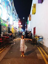 Portrait of happy girl with arms outstretched standing on street in city at night