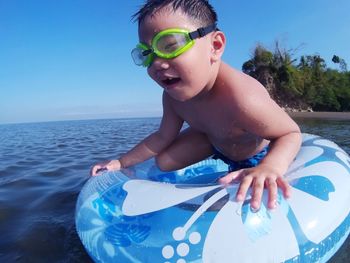 Boy wearing swimming goggles in inflatable ring over sea against clear blue sky