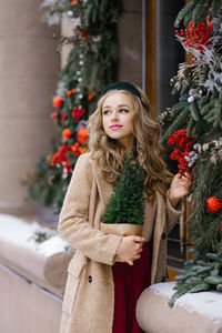 A young woman walking through the city's snow-covered streets holds a potted christmas tree 