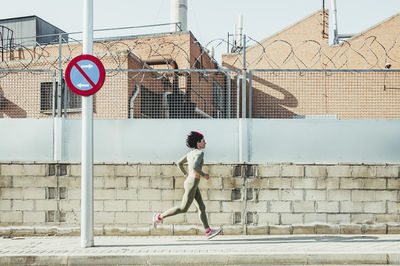 Woman in sports clothing running on footpath by brick wall