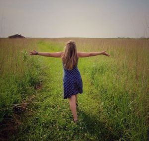 Rear view of woman with arms outstretched standing on grassy field against sky