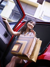 Portrait of smiling young woman holding shopping bags while getting out from car