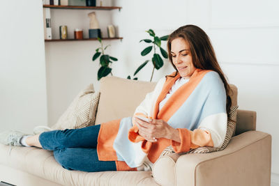 Young redhead woman sitting on the sofa in the living room uses the phone to communicate.