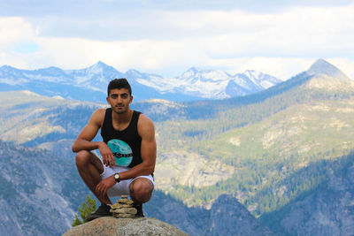 Portrait of young man crouching on cliff with mountains in background