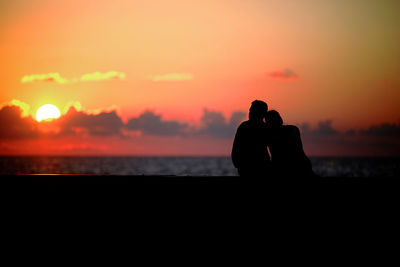 Silhouette couple sitting on railing at promenade against orange sky