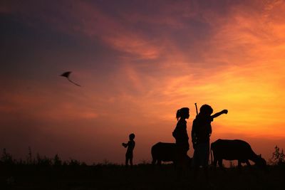 Silhouette kids and buffalos standing against orange sky during sunset