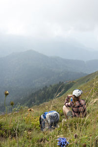 Young woman sitting on meadow in mountain valley next to backpack drinking water  on hike landscape 