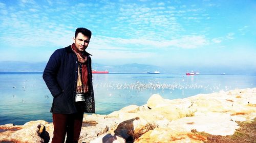 Portrait of young man standing on rock at beach against sky