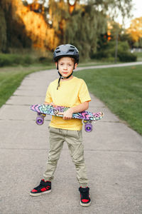 Happy caucasian boy in grey helmet standing with skateboard on road in park on summer day. 