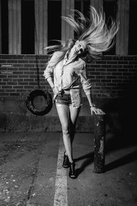 Full length of woman with tousled hair standing by bollard against wall